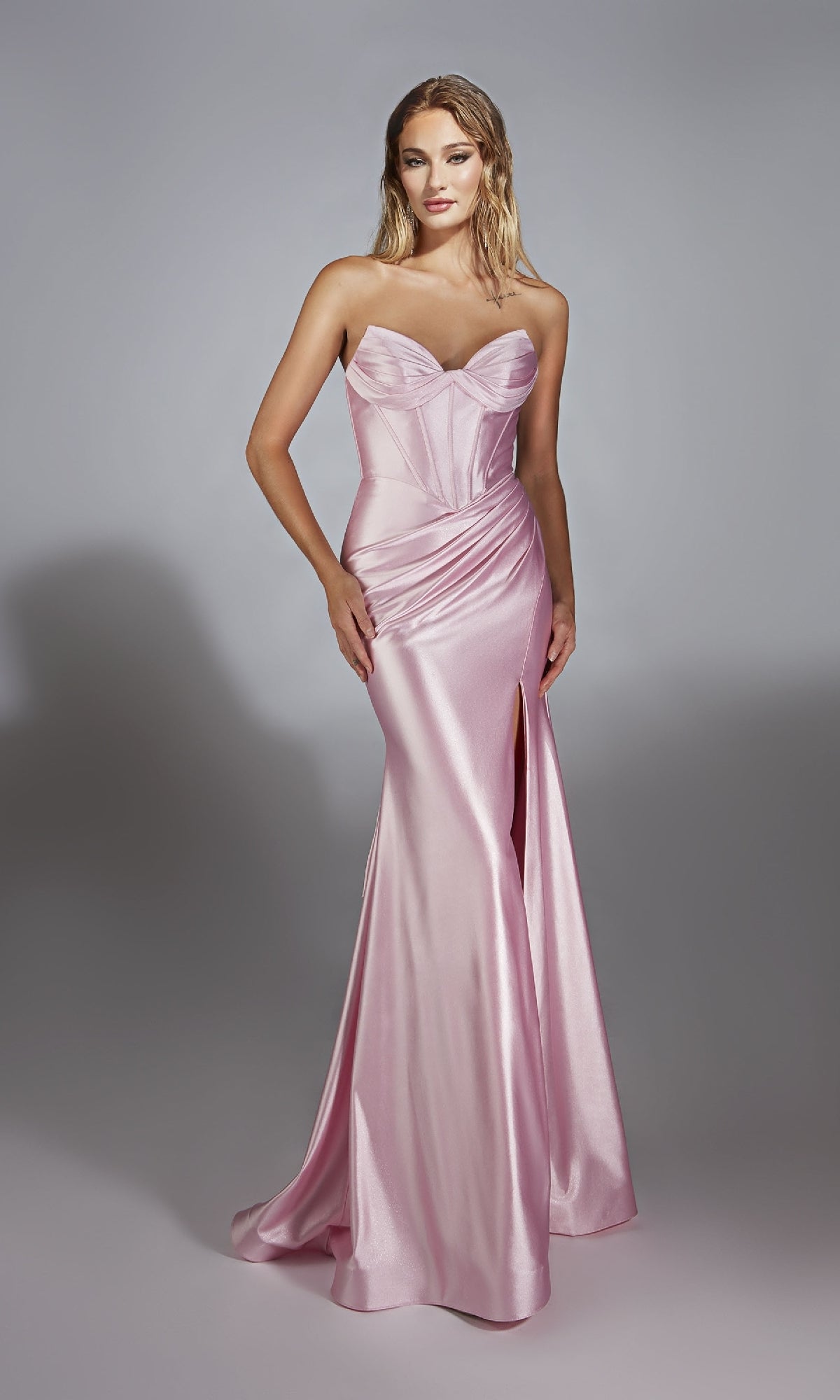 A woman wears the Alyce 61733 Strapless Long Corset Prom Dress in pale pink satin, featuring a fitted silhouette, sweetheart neckline, and high slit, standing against a plain gray background.